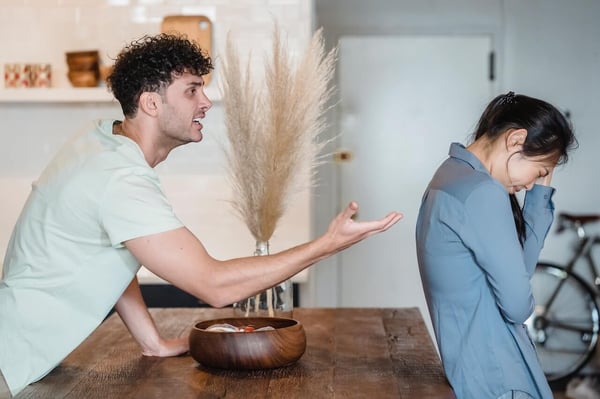 The image shows a white man and an Asian woman in an argument, standing on opposite sides of a counter. The man is frowning and gesturing toward the woman with his arm. The woman is flinching and has her back turned toward him. She is grabbing her face with her hand.