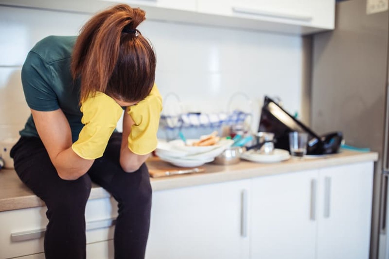 A woman wearing yellow cleaning gloves sits on a counter with her head in her hands. Dishes are piled on the counter behind her.