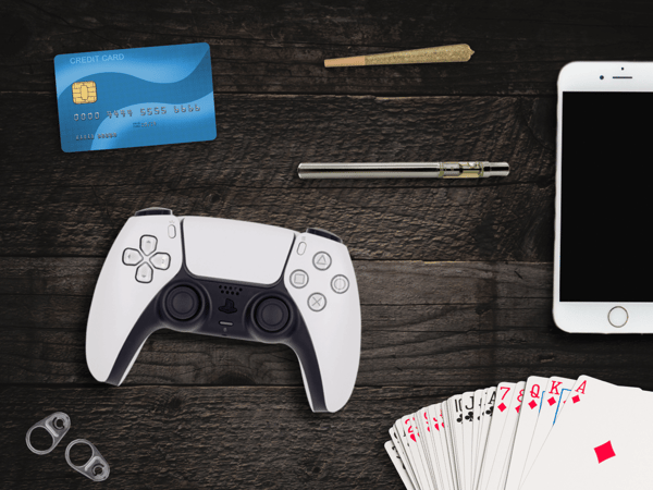 The image shows a credit card, a game controller, tabs from drink cans, a fanned-out deck of cards, a vape pen, a rolled joint, and a smartphone.