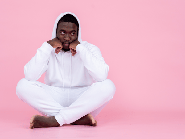 man in a white jump suit sits in front of a pink backdrop with his head resting on his hand.
