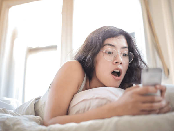 a woman with glasses lying on her bed and looking at her phone with a surprised expression.