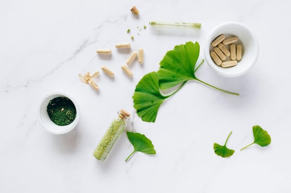 ADHD nootropic pills on a white marble background with green ginkgo leaves of different sizes dotted in between. There are two small white ramekins, one holding more pills, the other a dark green powder.