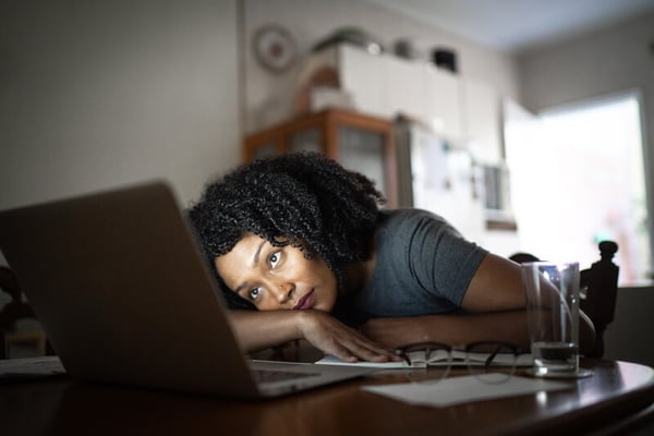 Woman with brown hair sits in dark room in front of an open laptop. She looks uninterested as she leans against the table.