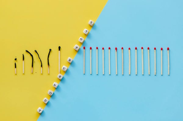 A colorful background divided diagonally by lettered dice; the background is yellow on the left side, light blue on the right. The dice spell out the words: Stop burnout. The left side has 6 burnt matchsticks next to each other. On the right side of the divide there are 14 unburnt matchsticks lined up.