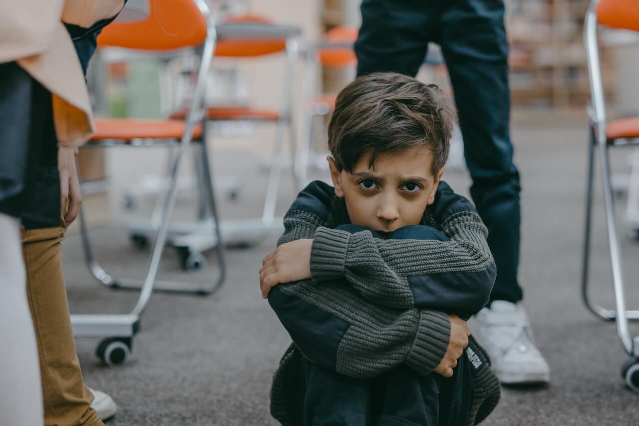 A child sitting on the floor in a classroom with his arms around his legs.